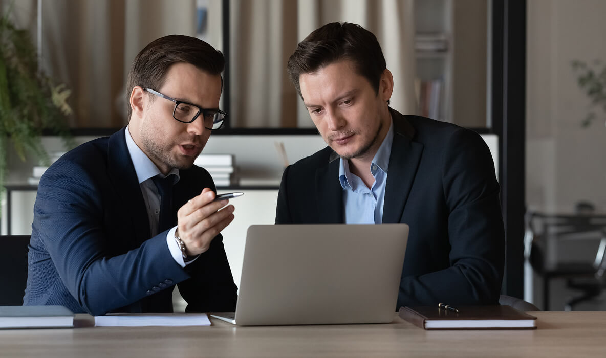 Dwóch mężczyzn w garniturach omawia coś przy laptopie podczas spotkania w biurze.